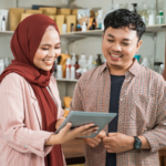 A smiling Muslim woman wearing a red hijab and a pink blazer holds a tablet while discussing something with a man in a plaid shirt. They are standing in a workspace with shelves filled with bottles and packaging, suggesting a business or financial planning setting.