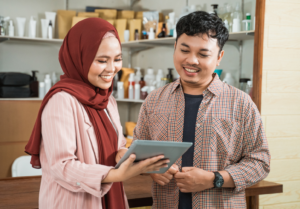 A smiling Muslim woman wearing a red hijab and a pink blazer holds a tablet while discussing something with a man in a plaid shirt. They are standing in a workspace with shelves filled with bottles and packaging, suggesting a business or financial planning setting.
