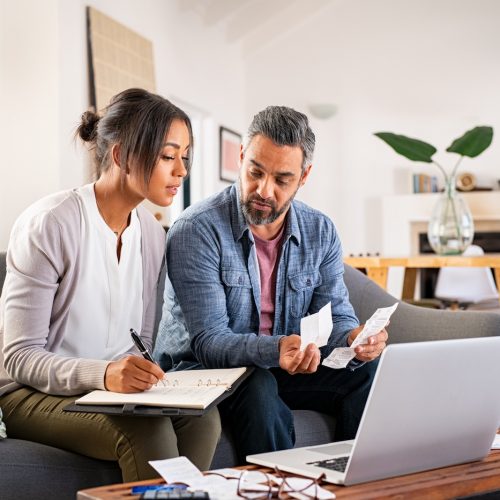 Mature couple calculating bills at home using laptop and calculator. Multiethnic couple working on computer while calculating finances sitting on couch. Mature indian man with african american woman at home analyzing their finance with documents.