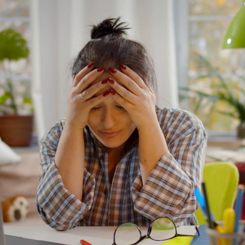 Tired mother with stress working on laptop at home with noisy children on background. Young exhausted woman putting off glasses and holding head sitting at desk with kids playing on background